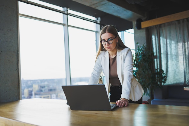 Smiling young woman in casual clothes and glasses wearing a white jacket and laughing while sitting at a table with a laptop while working Work in the office