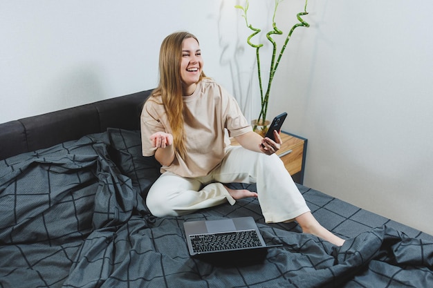 Smiling young woman in casual clothes smiling and working remotely sitting on bed with laptop during remote work Freelance work Work from home