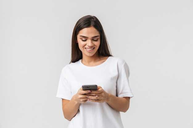 Smiling young woman casualy dressed standing isolated on white, using mobile phone