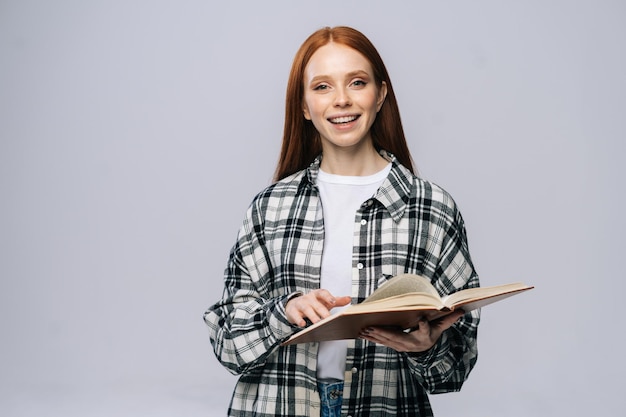 Smiling young woman college student turning pages of book while reading and looking at camera on gray isolated background Pretty redhead lady model emotionally showing facial expressions