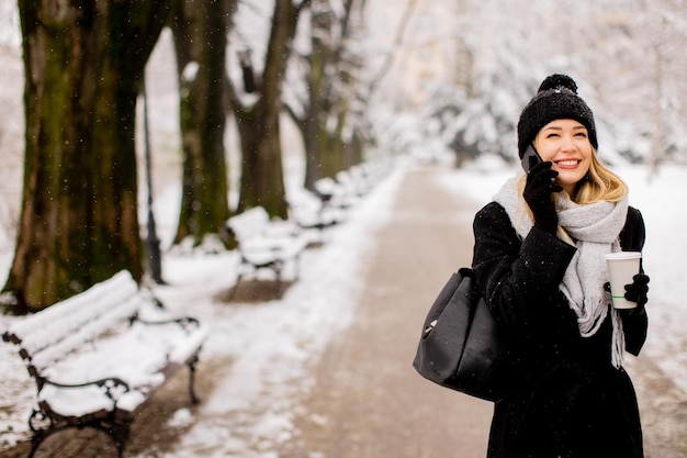 Smiling Young Woman in Cozy Clothing using mobile phone and holding coffee cup on winter day