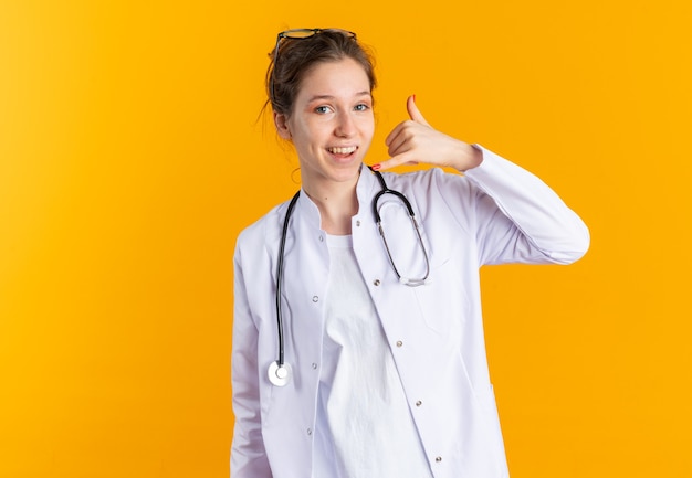 Smiling young woman in doctor uniform with stethoscope gesturing call me sign