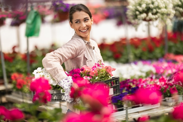 Smiling young woman florist is holding crate with beautiful flowers in a greenhouse.