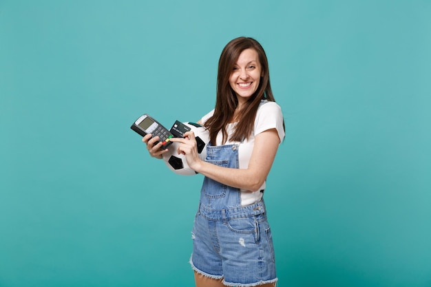 Smiling young woman football fan support favorite team with soccer ball, wireless modern bank payment terminal to process and acquire credit card payments isolated on blue turquoise wall background.