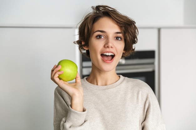 Smiling young woman holding green apple at the kitchen