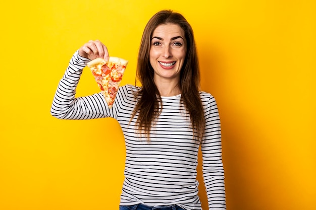 Smiling young woman holding a slice of hot fresh pizza on a yellow background