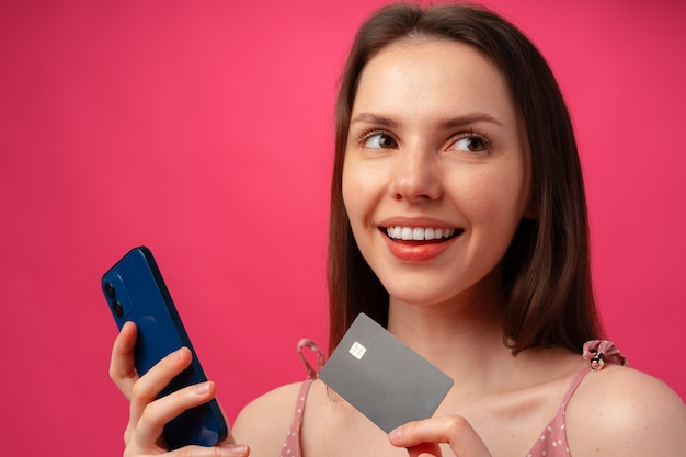 Smiling young woman holding smartphone and credit card against pink backgorund