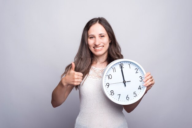 Smiling young woman holding white clock and showing thumb up.