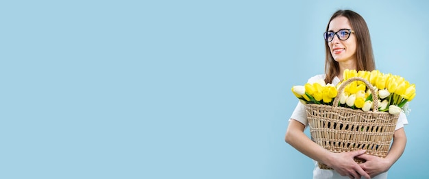 Smiling young woman holding yellow tulips in basket on blue background Banner