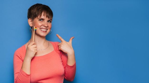 A smiling young woman holds a natural bamboo toothbrush, isolated on a blue background. The concept of oral hygiene. A place for your text.