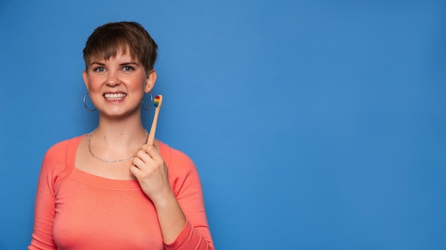 A smiling young woman holds a natural bamboo toothbrush isolated on a blue background The concept of oral hygiene A place for your text