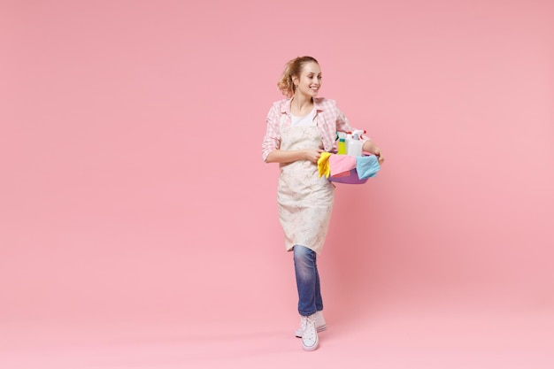 Smiling young woman housewife in apron hold basin with detergent bottles washing cleansers while doing housework isolated on pink wall background studio portrait. Housekeeping concept. Looking aside.