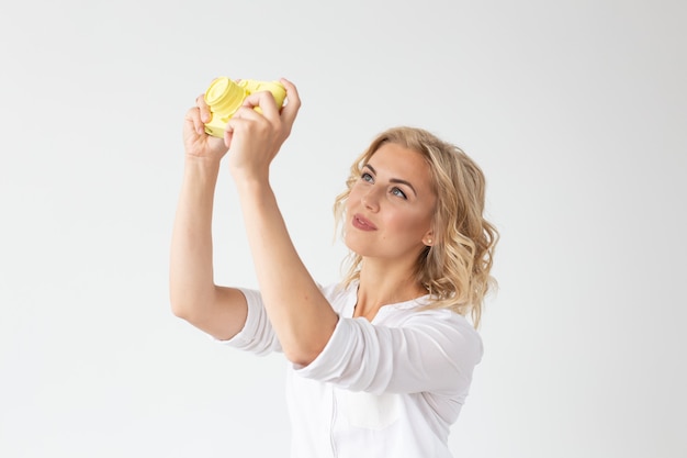 Smiling young woman is holding in her hands a yellow vintage camera model posing on a white wall. Concept of amateur and professional photography.