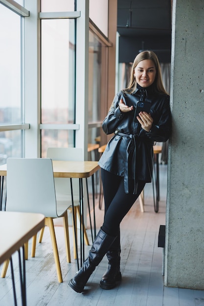 A smiling young woman is standing with a mobile phone in her hands and looking to the side while standing near a window reflecting sunlight Modern workspace Woman freelancer