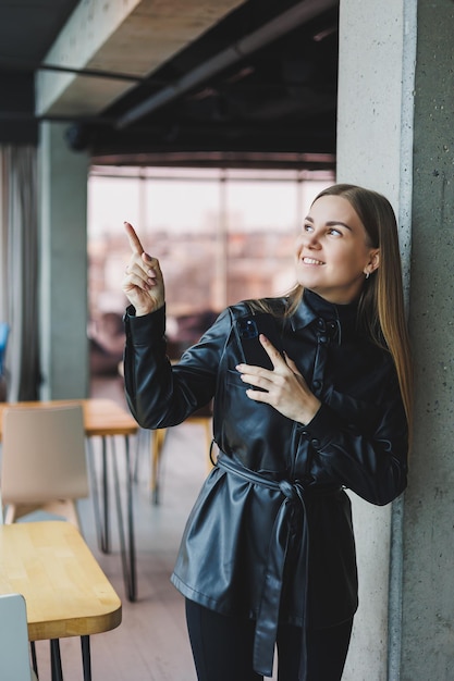 A smiling young woman is standing with a mobile phone in her hands and looking to the side while standing near a window reflecting sunlight Modern workspace Woman freelancer