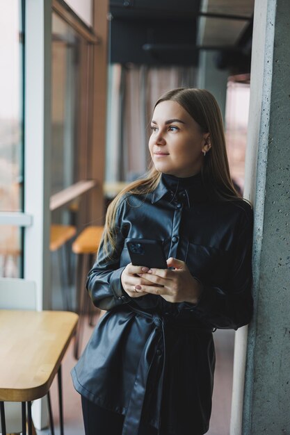 A smiling young woman is standing with a mobile phone in her hands and looking to the side while standing near a window reflecting sunlight Modern workspace Woman freelancer