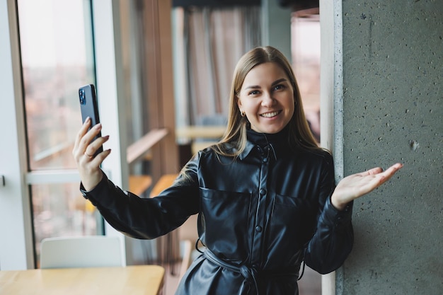 A smiling young woman is standing with a mobile phone in her hands and looking to the side while standing near a window reflecting sunlight Modern workspace Woman freelancer