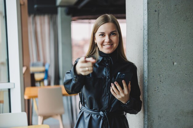 A smiling young woman is standing with a mobile phone in her hands and looking to the side while standing near a window reflecting sunlight Modern workspace Woman freelancer