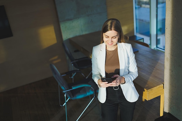 A smiling young woman in a jacket stands in the office and happily talks on the phone A young manager works in the office and manages the business remotely