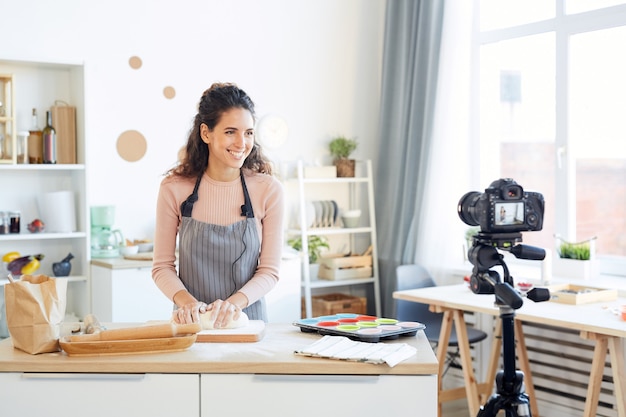 Smiling young woman kneading dough for cupcakes while shooting video for her food blog