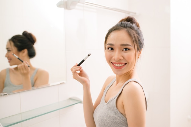 Smiling young woman looking to mirror doing makeup at home bathroom