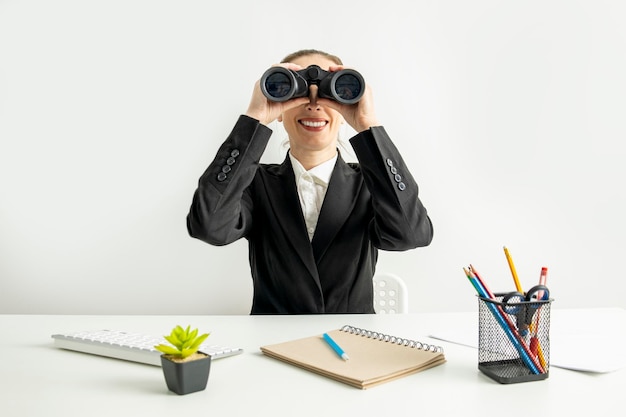 Smiling young woman looking through binoculars while sitting at desk at workplace