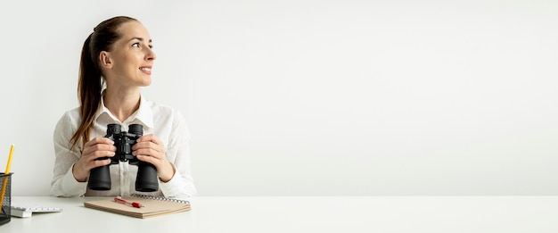 Smiling young woman looking through binoculars while sitting at office desk