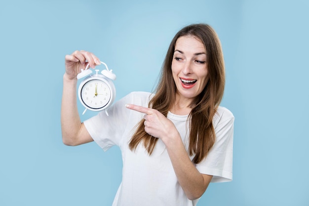 Smiling young woman pointing finger at alarm clock on blue background