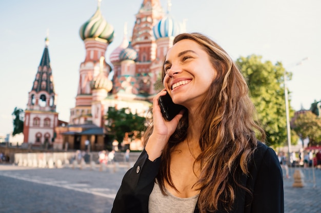 Smiling young woman portrait in Red Square talking at the phone, with Saint Basil's Cathedral in the background. Moscow, Russia.