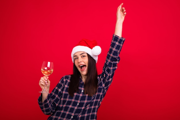 A smiling young woman in pyjamas wearing a Santa Claus hat holds a glass of wine on a red background New Year's Eve at home