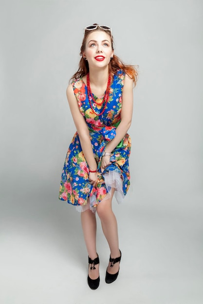 Smiling young woman Redhaired girl in a dress with a floral print red beads and sunglasses Gray background Vertical Full height