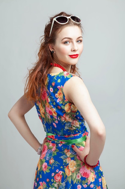 Smiling young woman Redhaired girl in a dress with a floral print red beads and sunglasses Gray background Vertical