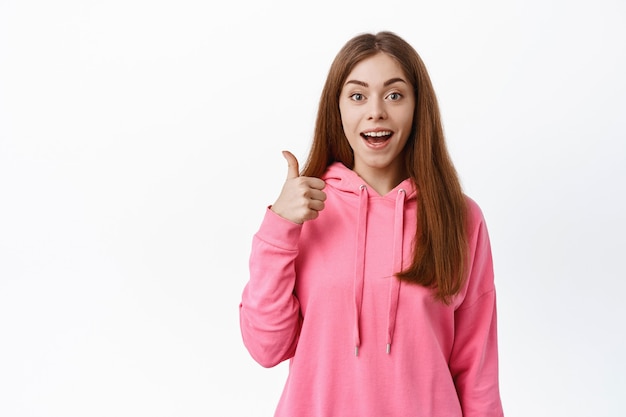 Smiling young woman say yes, agree with you, praise good choice, showing thumb up in approval, standing over white wall