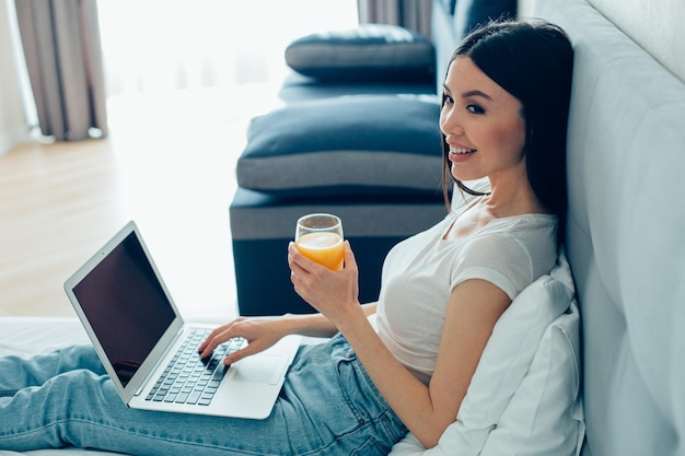 Smiling young woman sitting on a bed with a glass of orange juice and having a modern laptop on her laps