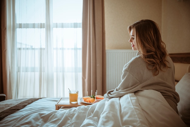 Smiling young woman sitting on bed with healthy glass of juice and fruits