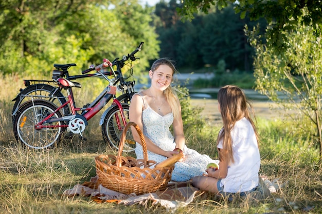 Smiling young woman sitting on blanket under big tree with her daughter