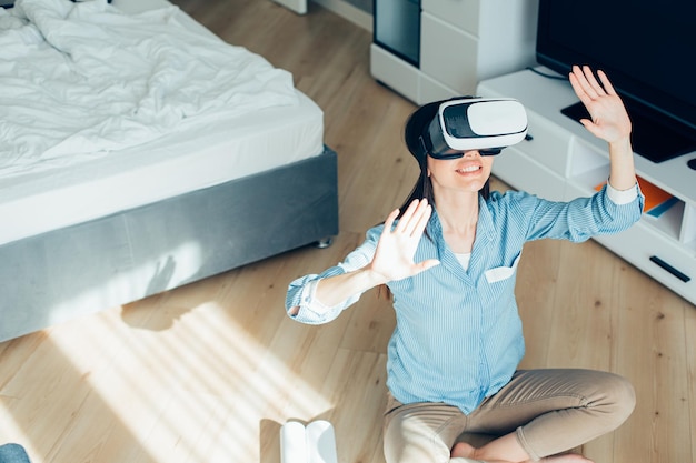 Smiling young woman sitting on the floor with hands up while using virtual reality glasses Website banner