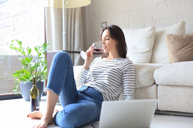 Smiling young woman sitting on floor with laptop computer and chating with friends, drinking wine.