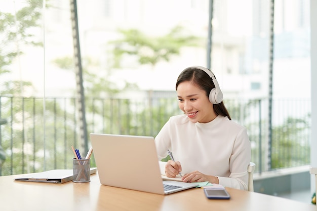 Smiling young woman sitting at the kitchen, using laptop computer for study