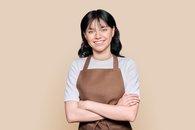 Smiling young woman staff in apron looking at camera on brown isolated background