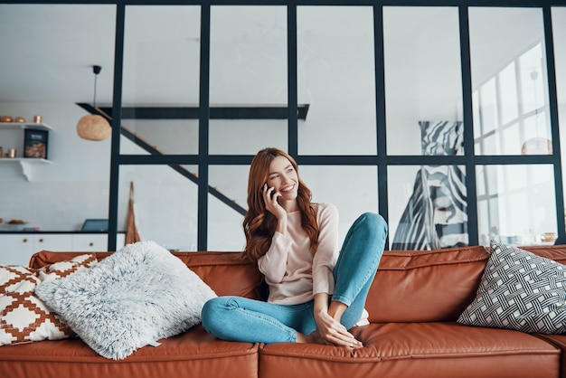 Photo smiling young woman taking on the phone while sitting on the sofa at home