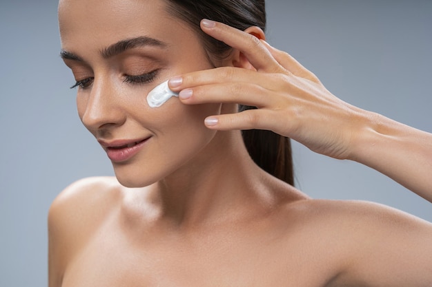 Smiling young woman using face cream on cheek