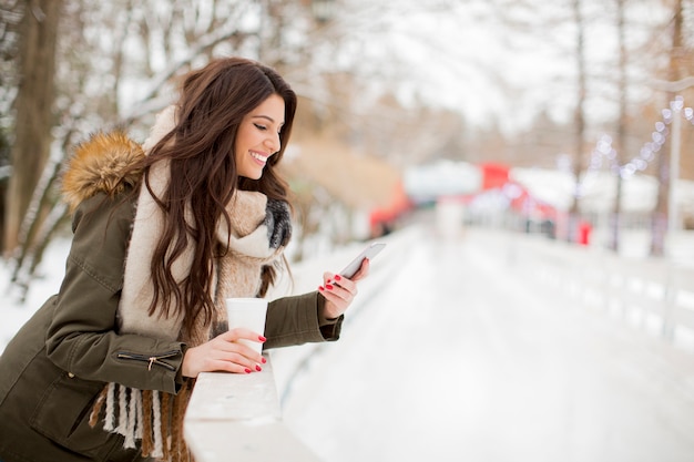 Smiling young woman using phone in park at cold winter day