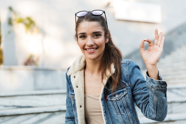 Smiling young woman wearing jacket sitting on a bench outdoors, showing ok