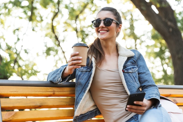 Smiling young woman wearing jacket and sunglasses sitting on a bench at the park, using mobile phone, drinking takeaway coffee