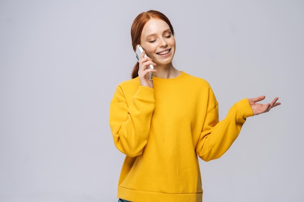 Smiling young woman wearing stylish yellow sweater talking by mobile phone on isolated white background