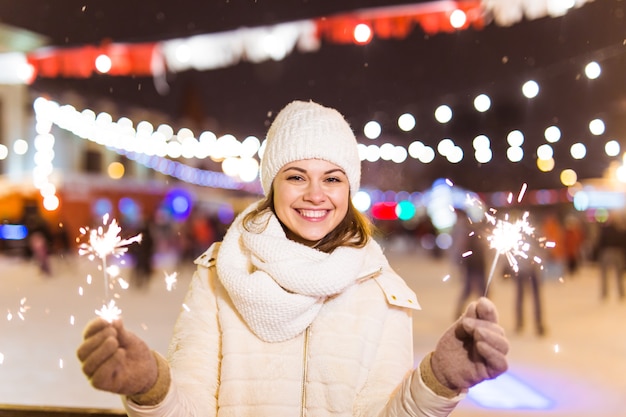 Smiling young woman wearing winter knitted clothes holding sparkler outdoors over snow background