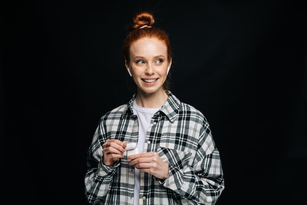 Smiling young woman wearing wireless earphones listening popular song on isolated black background