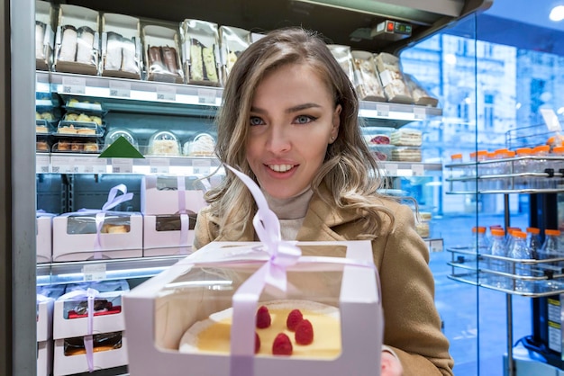 Smiling young woman with a cake in a beautiful package in a store Beautiful blonde in a beige coat Closeup