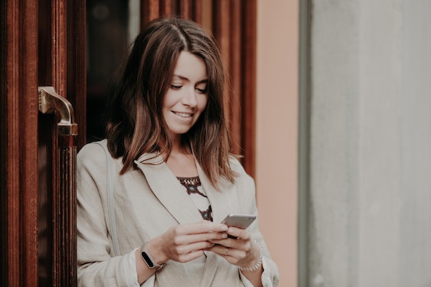 Smiling young woman with dark hair dressed in white elegant jacket holds modern smart phone stands near doors of office building messages with friends in social networks Technology concept